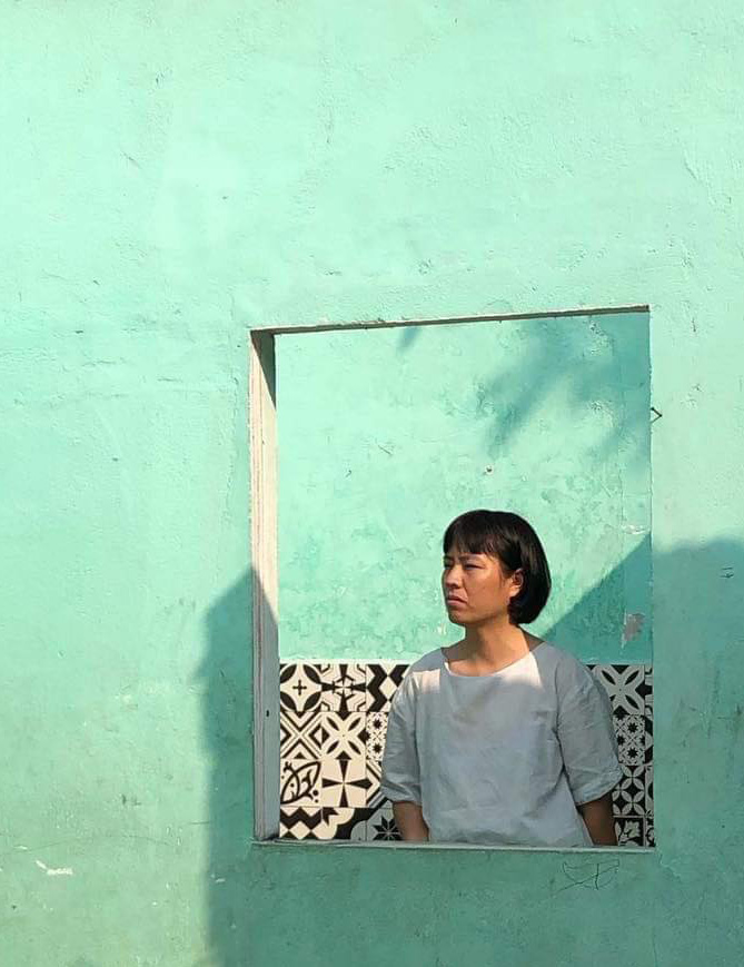 Person with a contemplative gaze framed in a square opening against a teal wall. // Person mit nachdenklichem Blick, gerahmt in einer quadratischen Öffnung vor einer türkisfarbenen Wand.